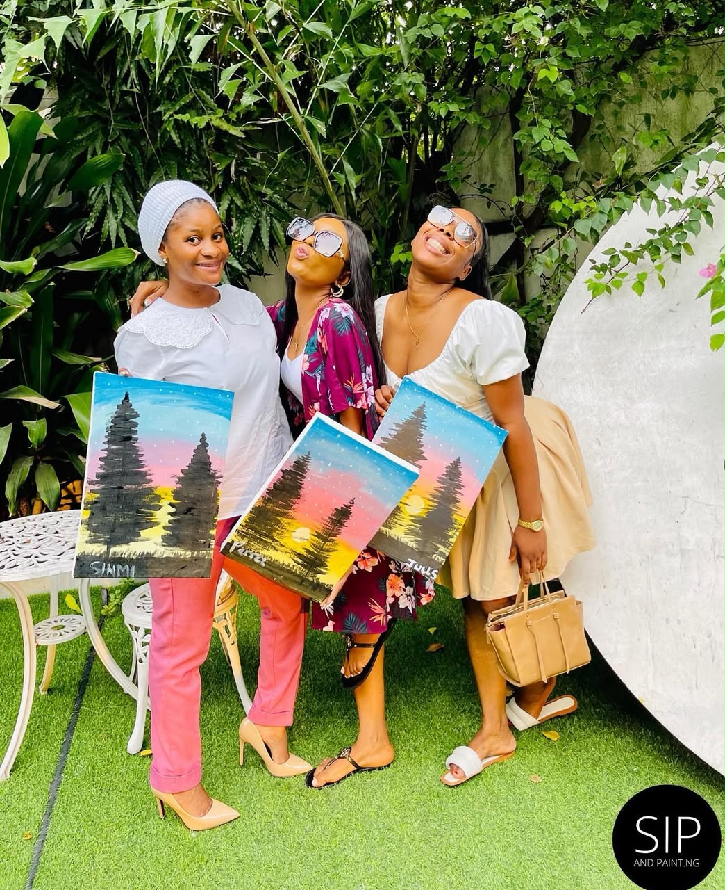 Three friends laughing in the garden with matching pine tree sunset paintings after a Sip and Paint Abuja session