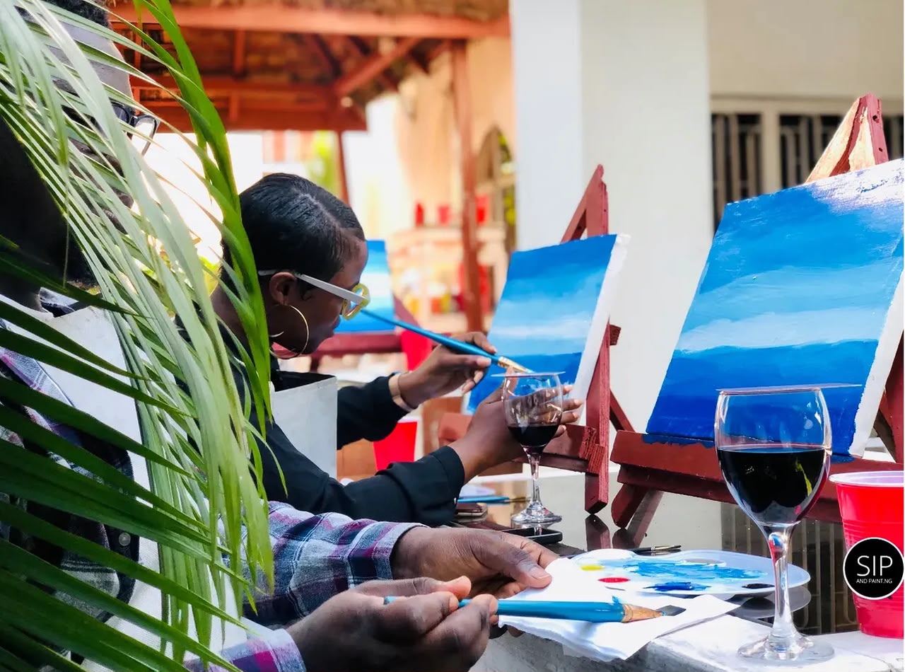 Close-up of guests painting a blue seascape with glasses of red wine at a Sip and Paint Abuja canvas session