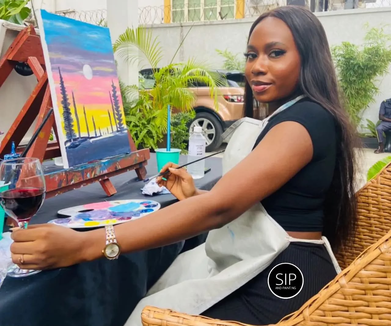 A guest painting a pink and yellow sunset with red wine at a Sip and Paint Abuja garden session