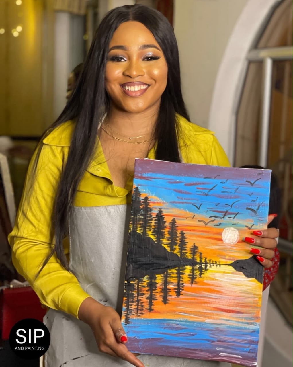 A guest in a yellow jacket smiling with her sunset forest lake painting at a Sip and Paint Abuja session