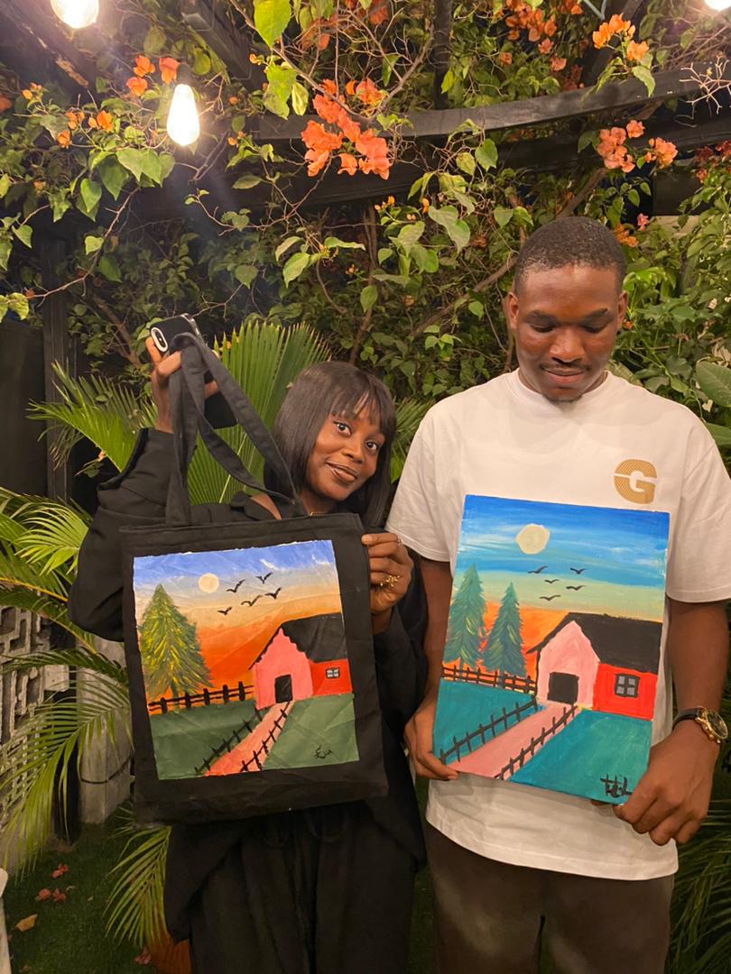 A couple standing under magenta bougainvillea in an Abuja garden, holding matching orange sunset paintings