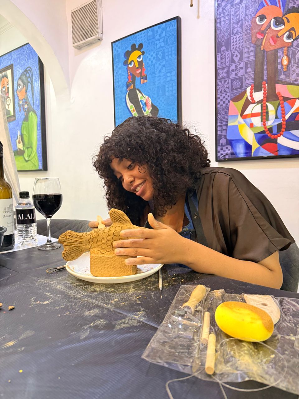 A guest smiling as she shapes a detailed clay fish sculpture on a plate at a pottery session, wine glass beside her