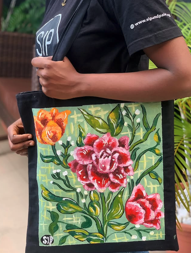 A guest in a Sip and Paint .NG tee holding a black canvas tote painted with vibrant pink, red and orange florals on a green background